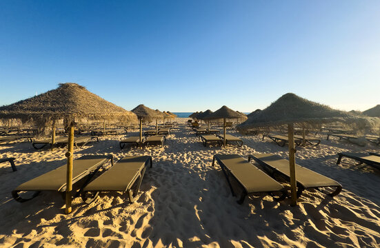 Beach at sunset with rows of sun loungers and straw umbrellas. The sky is painted in soft pastel shades of orange, pink and blue.