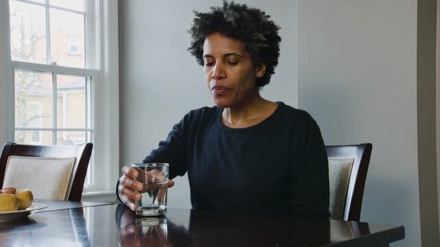 Thirsty woman in black lifting clear glass, sipping water, placing down at table by window, rolls