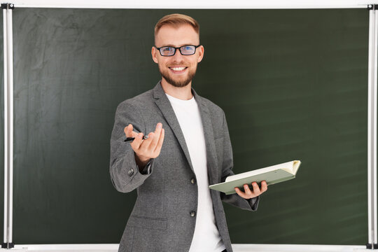 Smiling teacher in glasses and suit with book conducting lesson in front of chalkboard in classroom