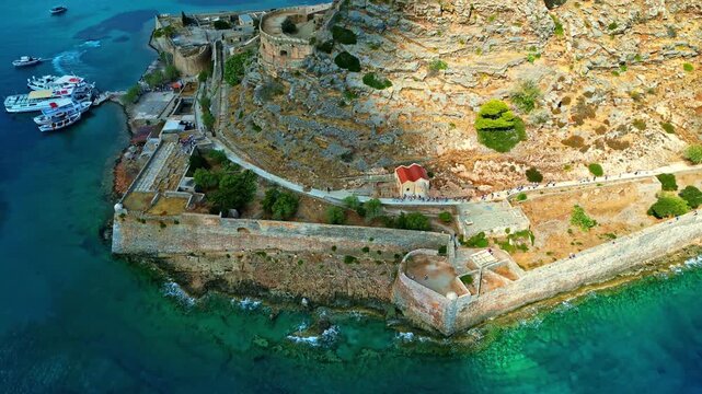 Aerial view of the historic Spinalonga fortress island with stone walls, a small church, and tourist boats docked in the clear blue sea of Greece.