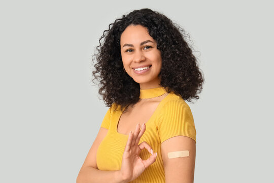 Beautiful young happy African-American woman with applied medical patch showing OK gesture on grey background