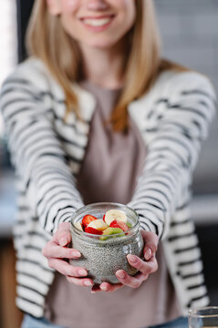 Woman holding a jar of chia pudding topped with fresh fruit toward the camera. Healthy breakfast, clean eating, nutrition and wellness concept with selective focus