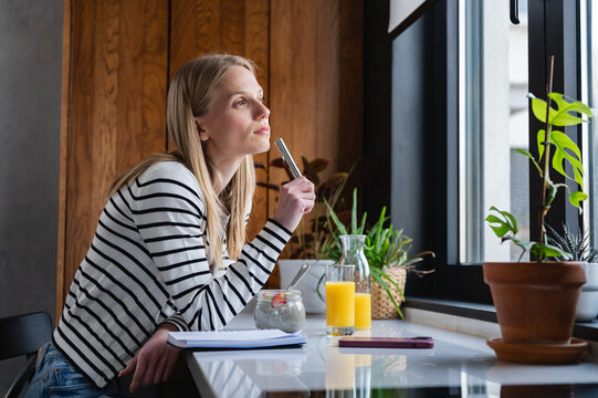 Thoughtful young woman holding a pen while looking through the window during breakfast in a modern kitchen. Planning, creativity, remote work and inspiration concept