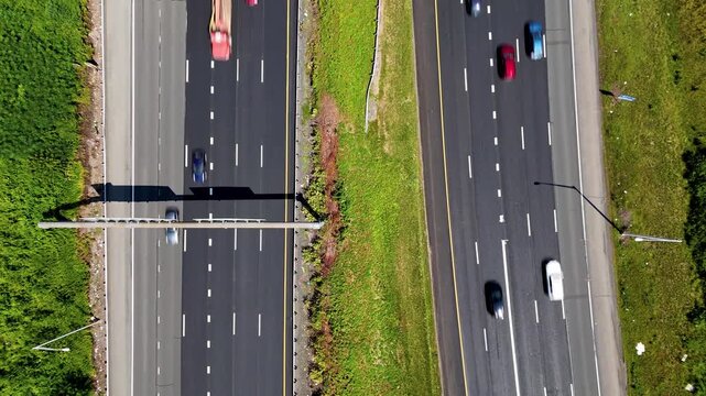 Top-down aerial drone pull away of traffic on a multilane highway with cars moving fast, grassy median, lane markings, and transport infrastructure. 
