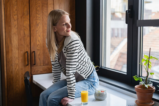 Young woman sitting on a kitchen countertop and looking through the window while enjoying a healthy morning. Cozy home, mindfulness and balanced lifestyle concept