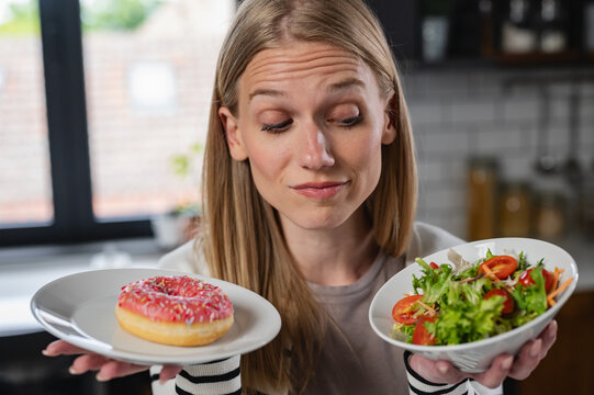 Woman weighing a healthy salad against a sweet donut in her kitchen. Concept of weight loss, mindful eating, healthy habits, nutrition and everyday food decisions