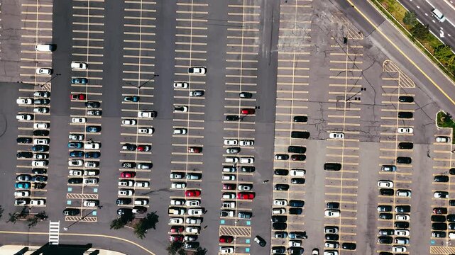 Overhead aerial view of a busy parking lot with parked cars arranged in marked spaces and adjacent roadways, representing urban transportation, infrastructure, and vehicle storage.