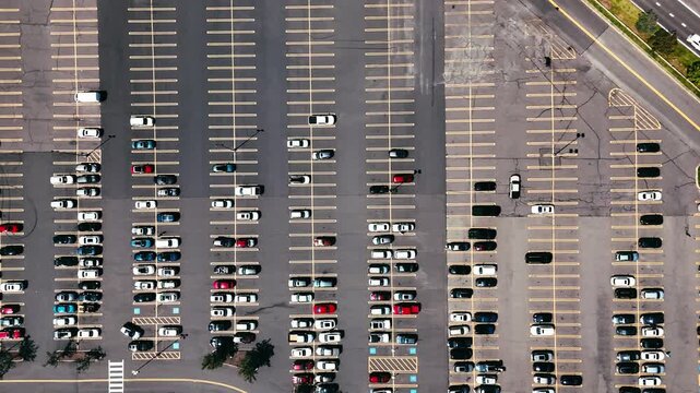 Top-down aerial view of cars parked in a large urban parking lot with marked spaces and nearby roads, representing transportation and city infrastructure.