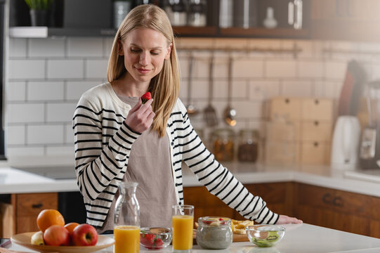 Happy woman savoring a fresh strawberry with a nutritious breakfast at home. Concept of healthy habits, mindful eating, modern living and everyday wellness