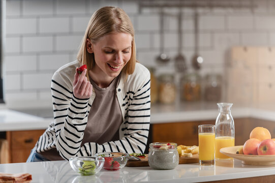 Young woman enjoying fresh strawberries and chia pudding during a healthy breakfast in a modern kitchen. Wellness, balanced nutrition, self-care and morning lifestyle concept