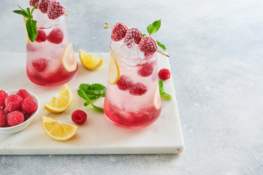 Refreshing raspberry iced drink with lemon slices, mint leaves, ice cubes and fresh berries on light table with copy space, summer beverage concept for mocktail recipe, lemonade menu, cafe promotion