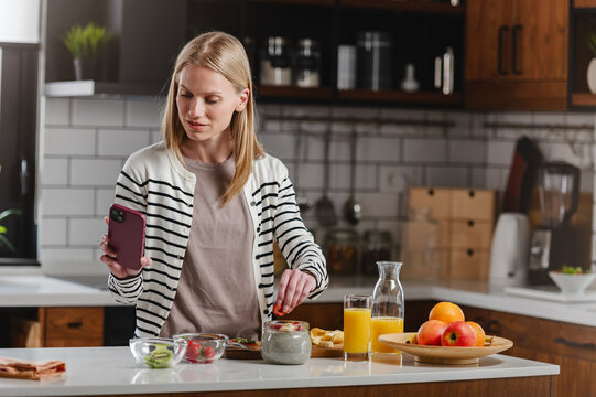 Woman using a smartphone while making a nutritious breakfast at home, illustrating modern routines, healthy eating, communication and work-life balance