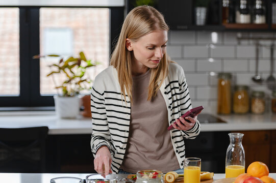 Young woman checking her smartphone while preparing a healthy breakfast with chia pudding, fresh fruit and orange juice in a modern kitchen, representing connected healthy living