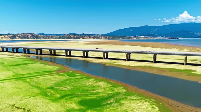 Aerial view of a long highway bridge spanning over dry lake bed with green moss meadow and water channel under clear blue sky.