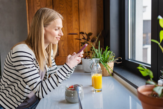 Young woman using smartphone while enjoying a quiet morning at home with fresh orange juice by the window. Cozy lifestyle, remote communication, social media, relaxation and wellness concept
