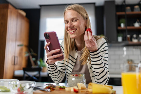 Smiling young woman enjoying chia pudding with fresh strawberries while using smartphone in a modern kitchen. Healthy breakfast, balanced lifestyle, wellness and home morning routine