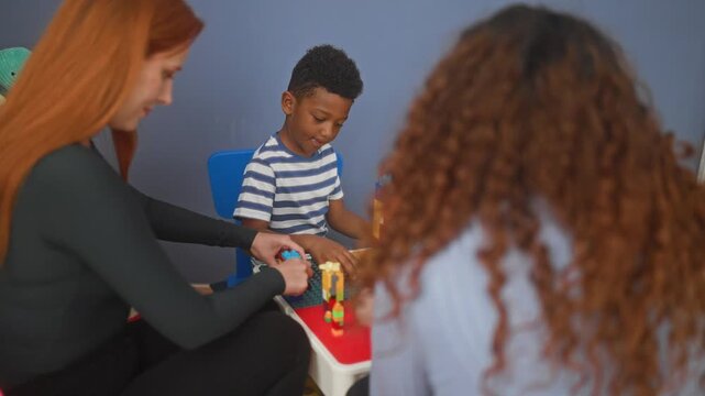 Woman guides boy stacking plastic building blocks at a classroom play table, hand on block while another woman watches; learning concentration.