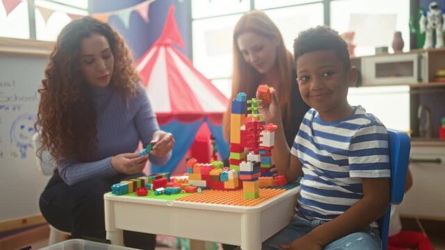 Boy holding a colorful plastic block at a preschool play table in a classroom building while teachers assemble bricks and he smiles; joyful teamwork.