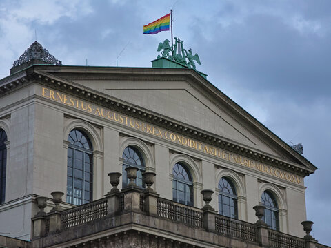 Hanover, Germany - 29 March 2026: View of the Opernhaus Hannover featuring neoclassical architecture, golden inscriptions, and a rainbow flag flying under a cloudy sky.
