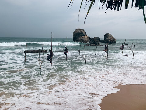 Sri Lanka - 25 April 2024: View of traditional stilt fishermen perched on wooden poles in the ocean with crashing waves and large rocks in the background.