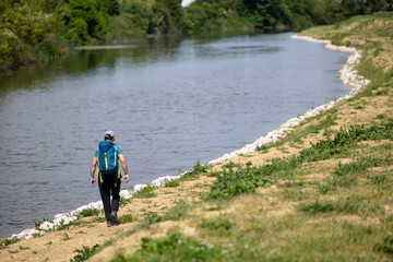 Fototapeta premium Solo Hiker With Blue Backpack Walking Along River Shore On A Sunny Day