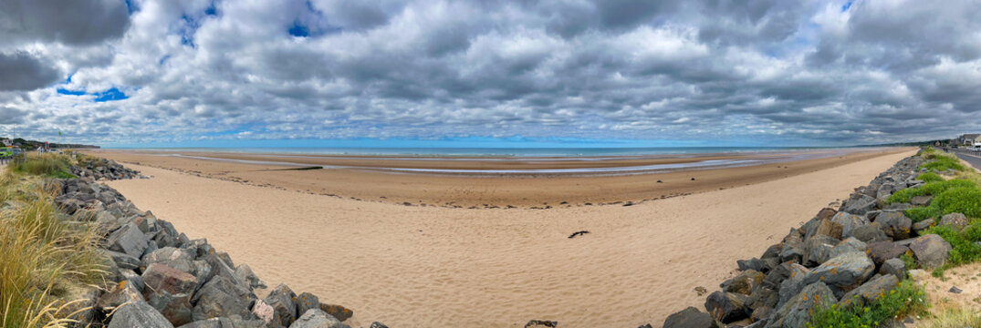 Panoramic view of Omaha Beach in Normandy, France. Historic WWII D-Day landing site with vast sandy shore, low tide, dramatic cloudy sky, protective rocks, and coastal landscape under overcast sky.