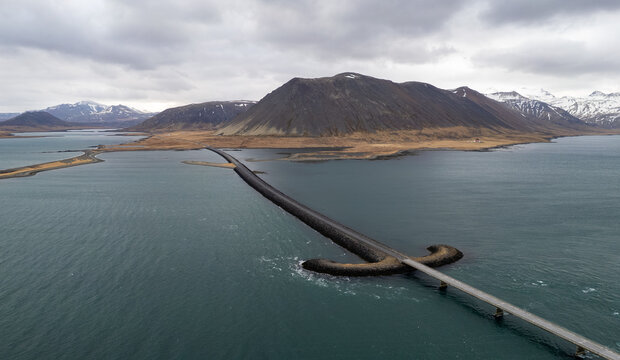Aerial view of a long causeway and kolgrafafjordur bridge stretching across an Icelandic fjord, framed by vast, rugged mountains under a dramatic, cloudy sky. Iceland