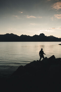 Holmstad, Norway - 30 August 2025: View of a man fishing from rocky shores at sunset with calm waters and distant mountains in the background.
