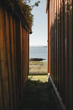 Straumsgard, Troms, Norway - 29 August 2025: View of the sea framed by two rustic wooden buildings with a white picket fence and calm water under a clear sky.