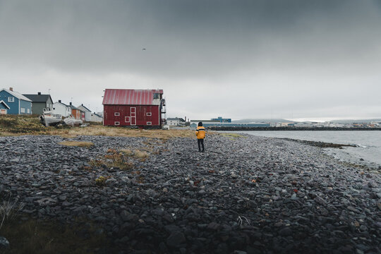 Vardo, Finnmark, Norway - 25 August 2025: View of a person in a yellow jacket standing on the rocky shoreline with a red wooden building and houses in Vardo, Finnmark, Norway.
