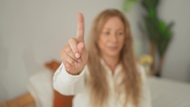 Woman points index finger at camera while seated on bed with pillows and a houseplant in building; calm focus.