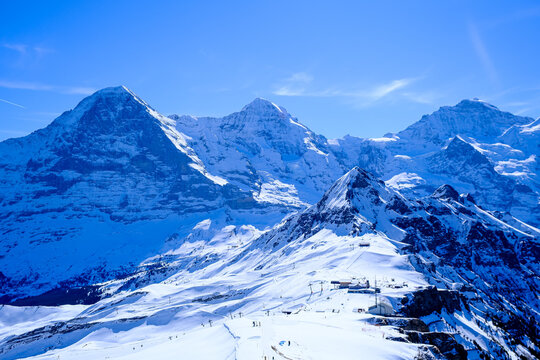A breathtaking, wide-angle winter landscape capturing the iconic snow-covered peaks of the Eiger, M&ouml;nch, and Jungfrau mountains in the Bernese Oberland region of the Swiss Alps. Shot on a clear, sunny
