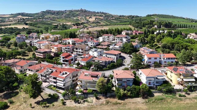 Panoramic aerial drone view of residential houses with orange roofs surrounded by green trees and rolling hills in Roseto degli Abruzzi Italy landscape.