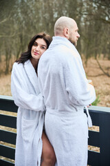 Obraz premium Woman hugging man from behind on terrace wearing bathrobes. Couple relaxing outdoors with forest view, morning calm, wellness retreat, cozy relationship and spa lifestyle concept