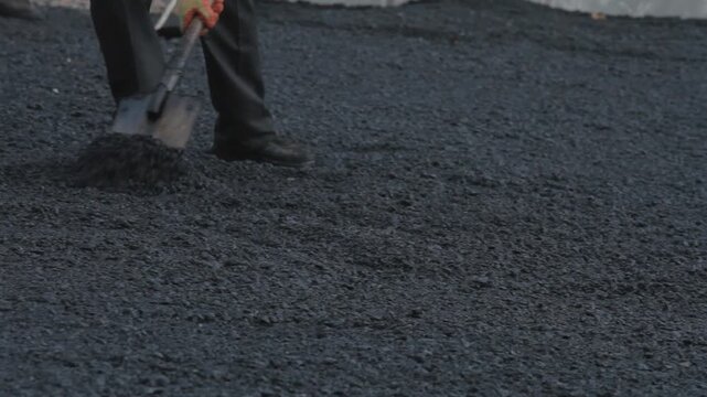 Close-up of a worker's legs using a shovel to spread fresh black asphalt on a road or driveway. Manual labor, road construction, and infrastructure repair.