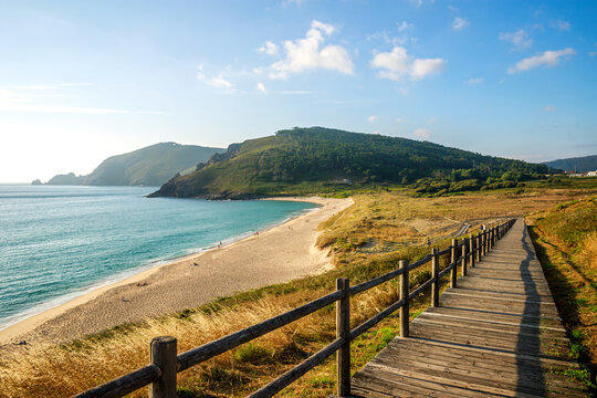 Wooden walkway leading to wild sandy beach, on background of green hills and blue sky, Fisterra, Spain