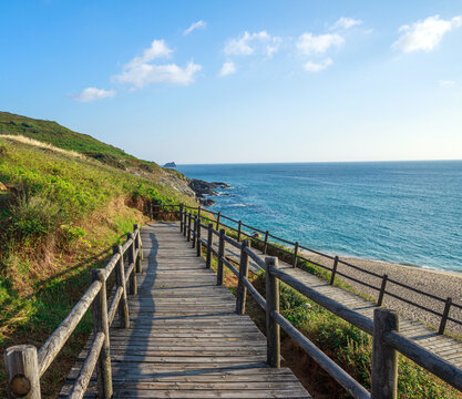 Wooden walkway leading to wild sandy beach, on background of green hills and blue sky, Fisterra, Spain