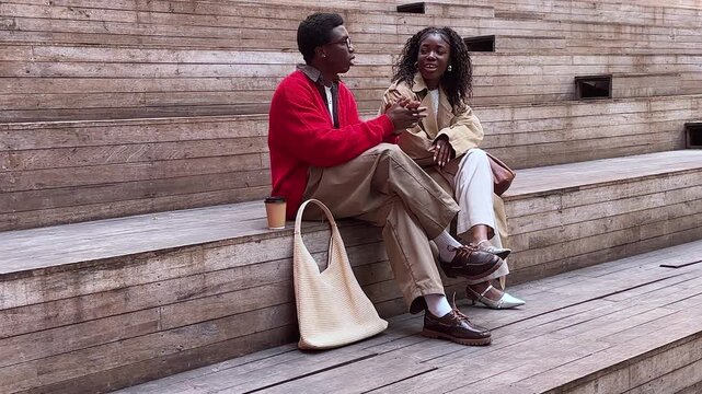 Positive african american friends students man and woman talking while sitting on steps outdoors, stylish young couple black male and female engaged in conversation. 