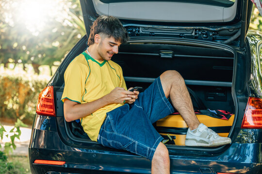 smiling young man in a summer outfit is sitting in the open trunk of a car, using his smartphone while traveling with a yellow suitcase on a sunny summer day