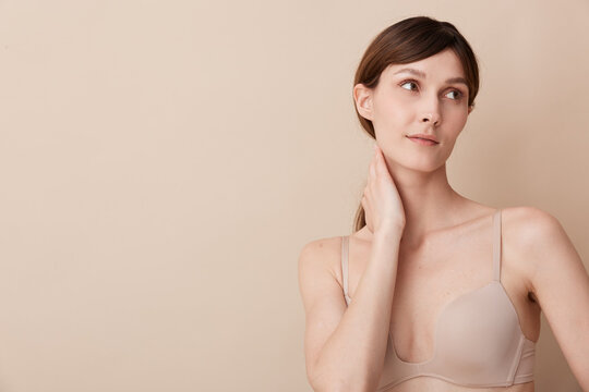A thoughtful woman stands in a soft beige studio, touching her neck with a calm expression. Subtle lighting highlights natural skin tones and a minimal wardrobe for a serene, intimate portrait.