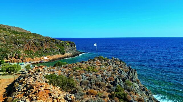 Aerial view of rocky coastline with a small beach and a white flag on the cliff overlooking the clear blue sea in Crete, Greece.