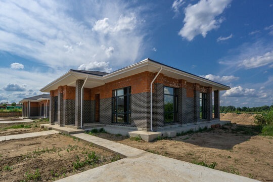 A new, stylish house stands among open land, showcasing a mix of brick and stone. The bright sky and fluffy clouds add to the peaceful rural atmosphere.