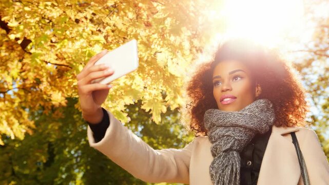 Young woman taking a selfie with a smartphone in a sunny autumn park with golden trees, wearing a warm coat and scarf during a fall afternoon