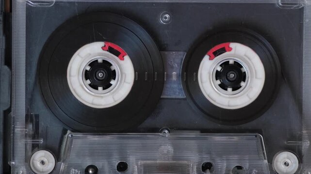Macro shot of a vintage audio cassette tape spinning inside a transparent plastic shell. Retro magnetic tape playing analog music with rotating spools