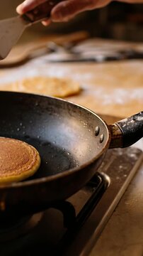 Vertical video: Sliding turner under pancake, hand with cuff lifting pancake on stove for browning