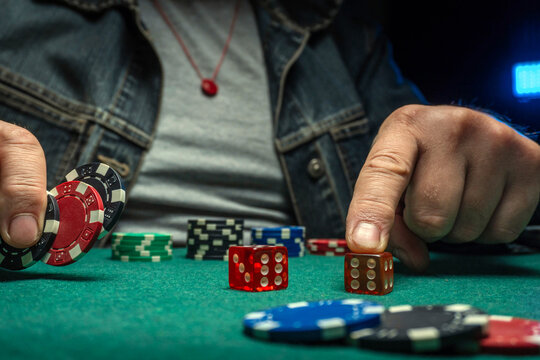 A man rolls dice and stacks poker chips on a green table. The setting is indoor with dim lighting at night. He appears focused on the game while sitting down