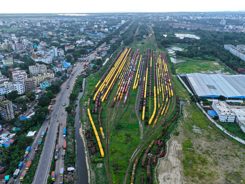 Aerial view of a large railway yard with numerous freight train cars lined up on parallel tracks next to a busy road and urban buildings in Chittagong, Chittagong Division, Bangladesh.