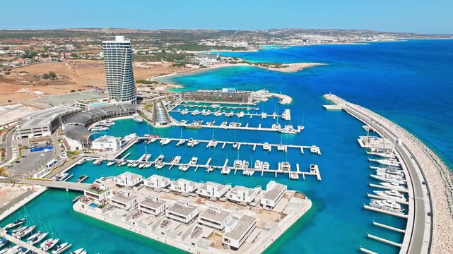 Drone view of Ayia Napa Marina and coastline in Cyprus. The tower, marina berths, harbour walls and bright blue sea form a striking coastal resort scene.