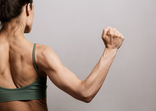 Young strong woman showing her muscles with visible veins, tendons, ligaments on her arm and hand. Sport fitness female showing biceps and strong upper back on grey studio background with copy space