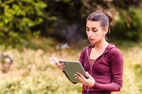 Young woman smoking cannabis joint outdoors using tablet